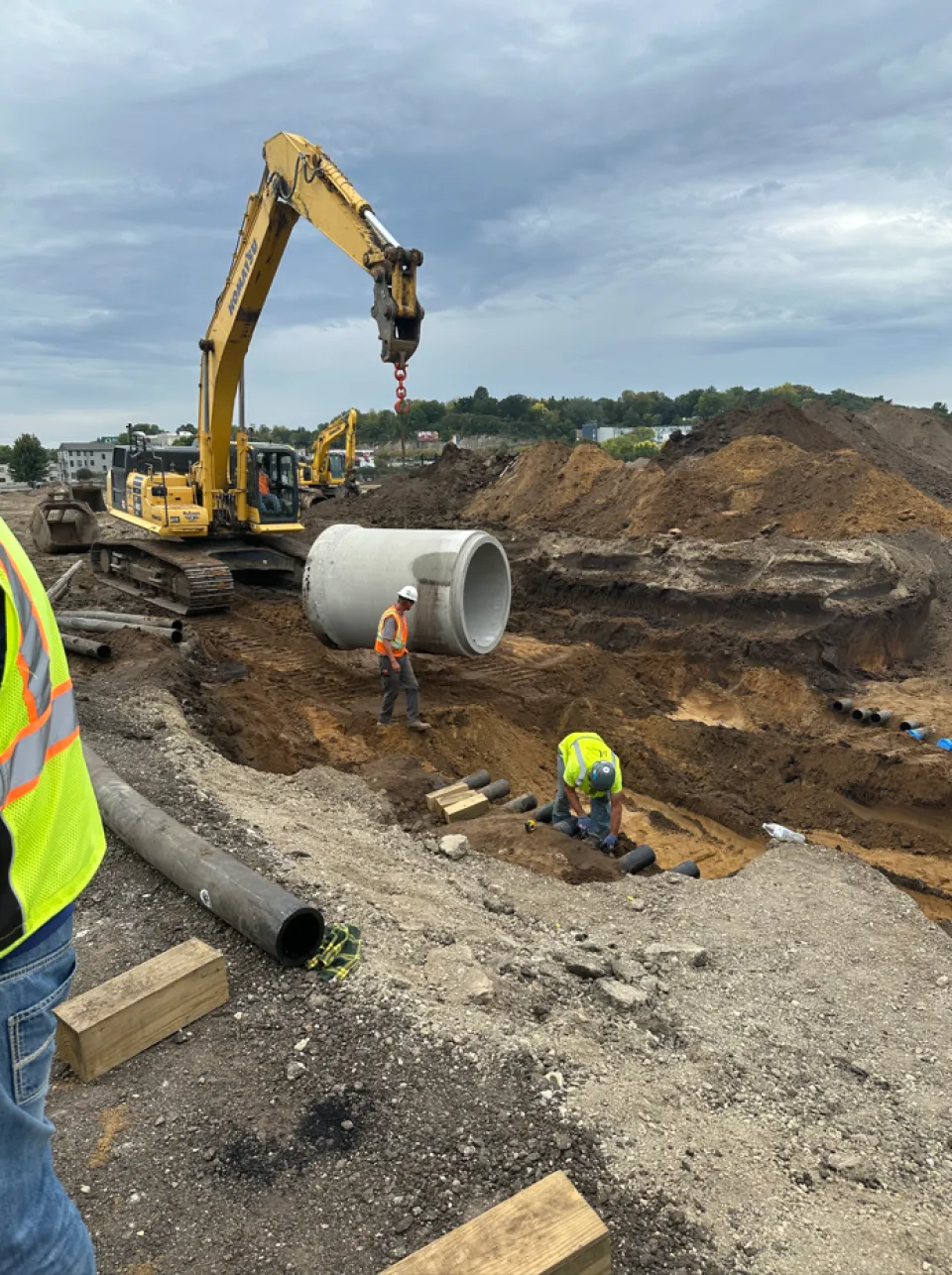 Construction workers and a large excavator at a work site, installing concrete pipes in a trench.