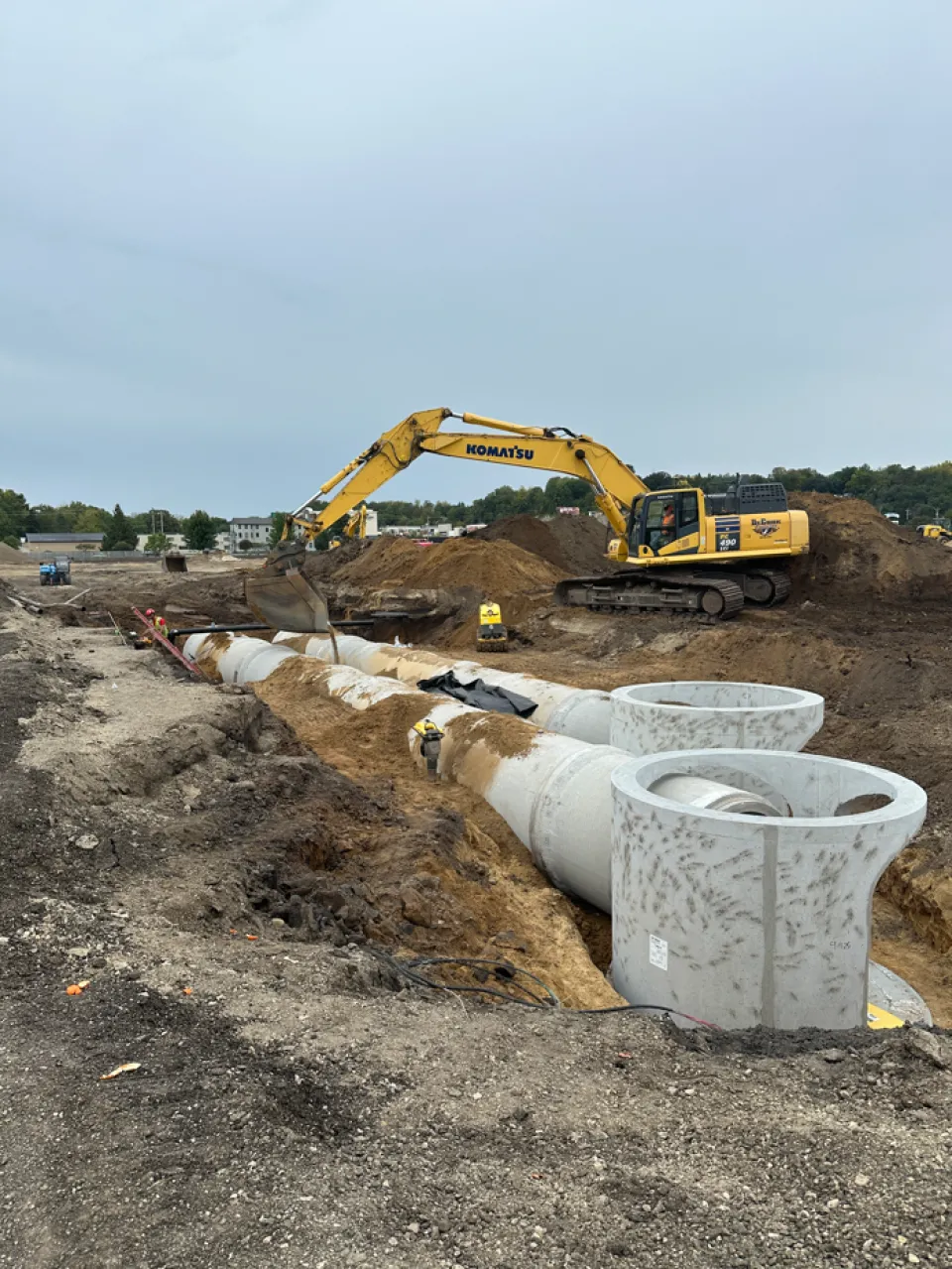 A construction site featuring a Komatsu excavator working near large concrete pipes laid in a trench, with a partially developed residential area in the background.