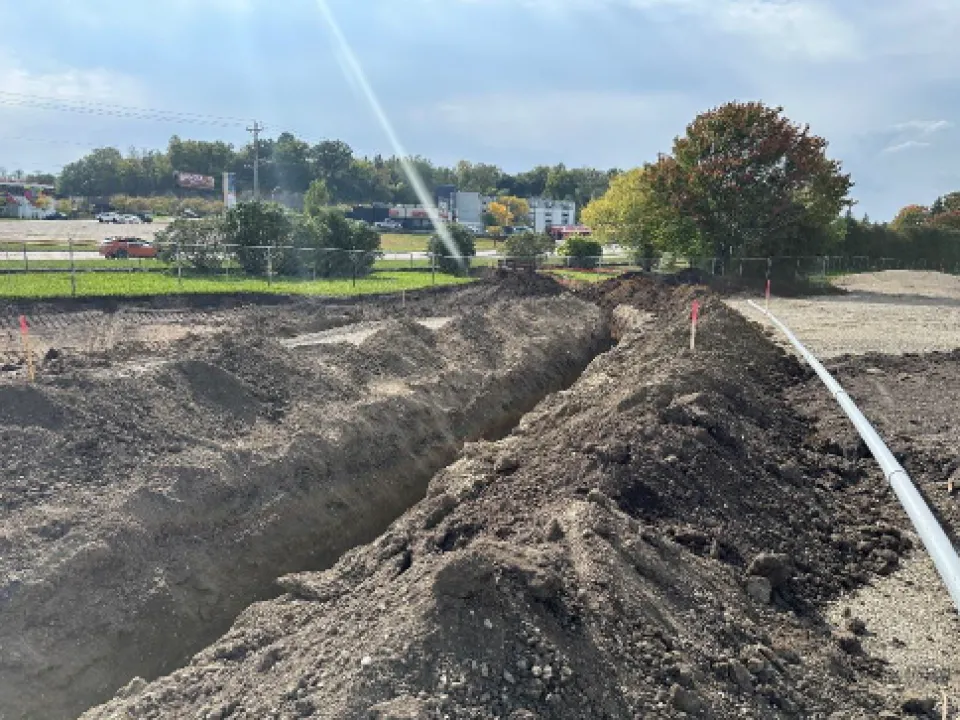 A construction site with a large dug trench and mounds of dirt, with a white pipe running alongside the trench. In the background, trees and cars passing by are visible under a clear sky.