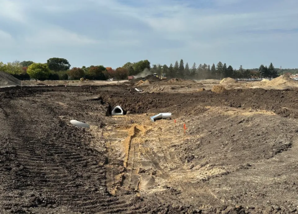 Large construction site with exposed earth, several large pipes visible on the ground, and construction equipment in the background under a clear sky.