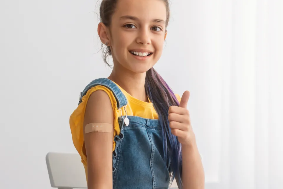 A girl sitting on a chair posing to show her band-aid after receiving a vaccine and showing a thumbs up.