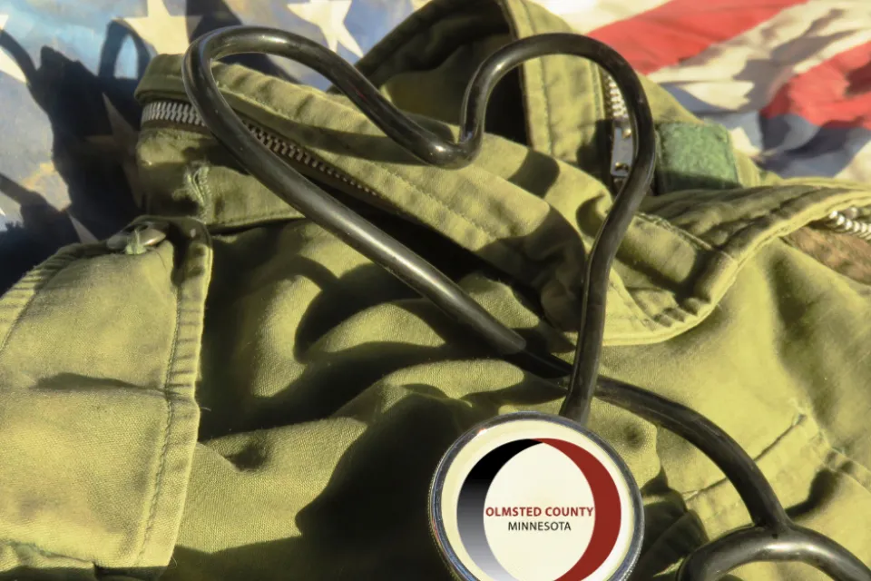 A stethoscope is placed on top of a green jacket, which is partially covering an American flag. The stethoscope features a badge labeled "Olmsted County Minnesota.