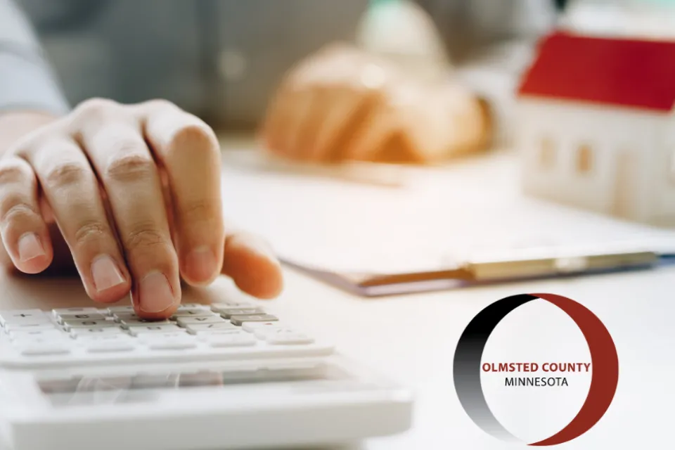 An individual is using a calculator with documents and a small model house on a desk, next to a logo of Olmsted County, Minnesota.